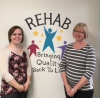 Two smiling women standing in front of a sign reading Rehab, Bringing quality back to life